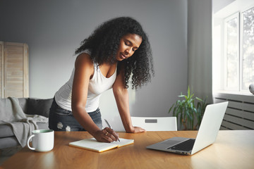 Busy day of modern African female who is standing by wooden desk in cozy room, writing down...