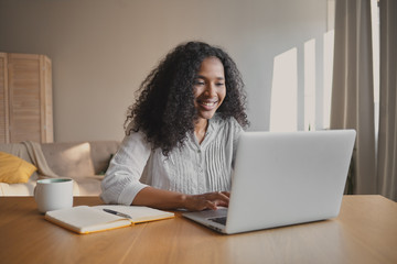 Cheerful young African American woman copywriter sitting in front of open laptop with mug and...