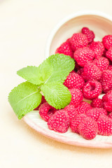 Ripe raspberries with green mint leaves in cup and saucer on pastel yellow background.