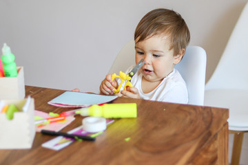 Little baby boy holding scissors in his hands and making handmade paper greeting card for mother. Fine motor skills development. DIY concept. Child safety.