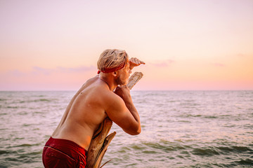A tanned man stands on the coast leaning on an old tree, looking for a ship on the horizon that can...