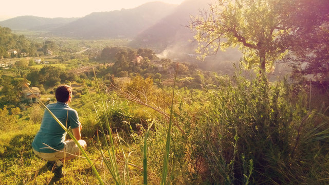 Hombre joven agachado observando pueblo de Bunyola, valle y monta&ntilde;as desde una colina con bosque y vegetaci&oacute;n verde en d&iacute;a soleado de primavera al atardecer en isla de Mallorca, Islas Baleares.
