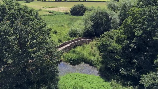 Aerial perspective view, forward move and stop. Drone fly towards old small sandstone roman bridge and grass, overgrown water pond in english countryside.