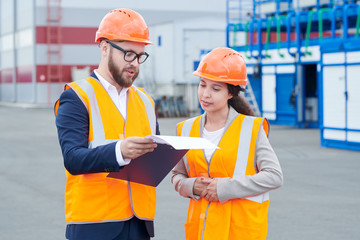 Waist up portrait of two modern factory workers wearing hardhats discussing production over clipboard outdoors, copy space