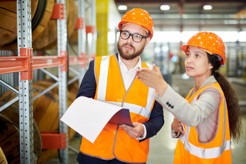 Waist up portrait of female factory worker talking to bearded businessman wearing hardhat and...