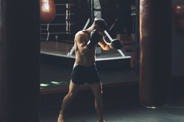 Topless athlete is training defense and attacks in sport center. He is kicking punching bag while...