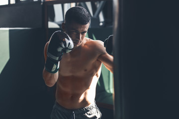 Shirtless shredded guy is having combat training in gym. He is kicking punching bag while wearing...