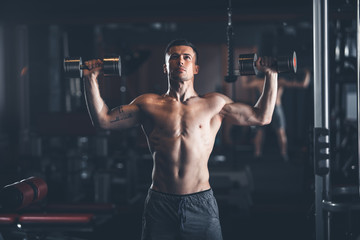 Fototapeta na wymiar Shapely topless guy is standing and lifting dumbbells in gym. He is pressing equipment up with effort. Strong male training concept
