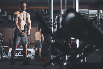 Fototapeta na wymiar Focus on smiling topless athlete while exercising in sport club. He is standing near machine and pulling down cable with weight for triceps relief. Copy space in right side