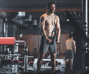 Fototapeta na wymiar Motivated topless man is having strength workout with different equipment. He is standing near machine and pulling down handle while exercising arms. Sportsman is working with weights