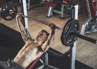 Top view of shapely topless man exercising with sport machines. He is lying on bench and lifting heavy barbell. Sportsman is working on chest muscles