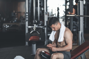 Tranquil strong guy with towel is sitting on bench of gym machine. He is resting between training with barbell and surfing internet with his smartphone. Copy space in left side