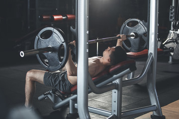 Ripped athlete is doing chest press while using gym machine. He is lying on bench and holding...