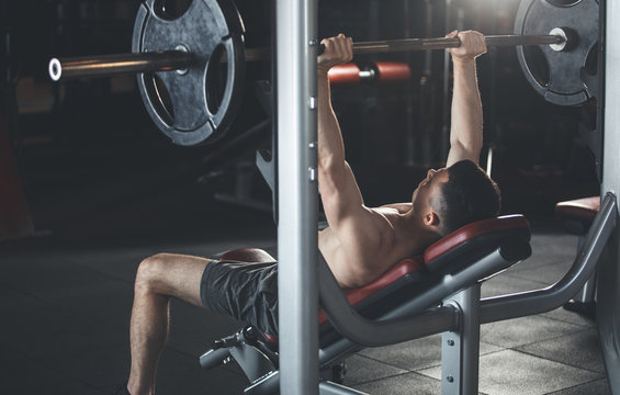Shredded sportsman is working on chest muscles while using machine and equipment. He is lying on bench and lifting heavy weight by doing press ups. Strong male concept