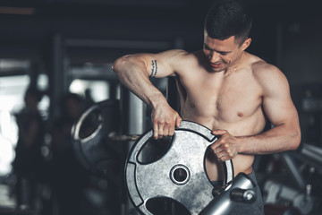 Fototapeta na wymiar Positive shirtless man is exercising in gym. He is using heavy barbell for improving muscles. Male is standing near machine and putting plate on equipment