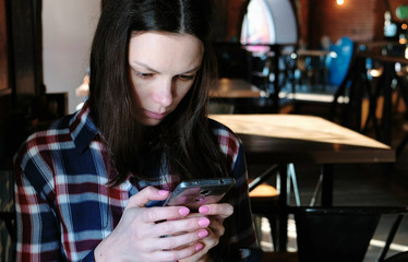 Upset woman sends a message or use internet in the phone sitting in a cafe. Dressed in a plaid shirt.