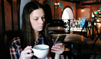 Brunette woman in checkered shirt drinking tea from a large cup and looking at phone.