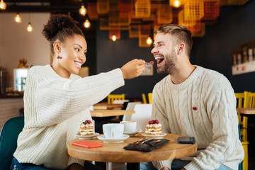 Piece of cake. Caring beautiful curly girlfriend giving piece of cake her husband while sitting in coffee shop