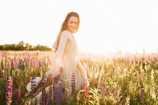 Beautiful dark-haired woman in a summer dress in a field of blooming lupine flowers with a straw hat in hands