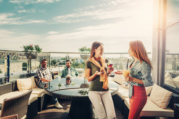 Long time no see. Smiling lovely women enjoying conversation at rooftop bar. Handsome bearded gentlemen on background