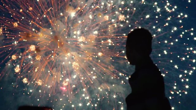 Silhouette of boy who watch the fireworks in the night sky. Bright and colorful lights in the sky. A beautiful sight is watched by the guy. Red, green and orange lights in the blue sky