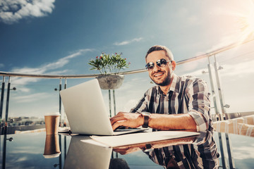 Feeling joyful. Handsome writer looking at camera with smile while sitting at the table with cup of coffee and notebook computer