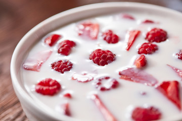Close-up view of red ripe raspberry, pieces of strawberry with fresh milk in a bowl on a wooden table.