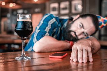 Cold dark beer. Bearded dark-haired man looking at glass of nice cold dark beer while sitting alone in the bar