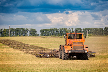 An orange modern tractor plows the earth in.  a golden field of wheat on a summer day, in the sky a cumulus cloud, in the background a forest.