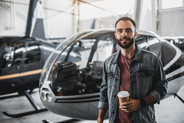 Portrait of cheerful bearded man tasting appetizing cup of coffee while standing near contemporary helicopters inside special garage for them. He wearing casual clothes and modern watch