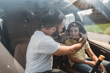 Happy male pilot putting headset on cheerful kid. They locating inside of rotorcraft during communication
