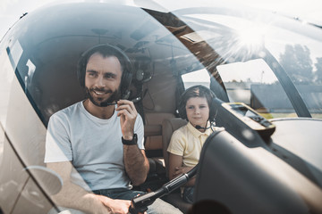 Portrait of happy unshaven pilot and smiling child sitting in cabin of contemporary helicopter. They wearing headsets. Glad male talking in microphone