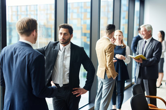 Two young elegant men in suits standing by window of modern conference hall while discussing details of report