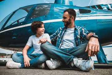 Side view outgoing boy speaking with smiling unshaven male. They sitting on ground near rotor plane...