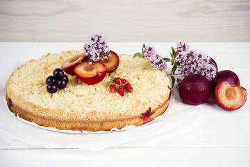 homemade pie with plums decorated with currants, strawberries and flowers on a white wooden table