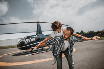 Portrait of beaming bearded man having fun with laughing little boy. He keeping him on back during game on air-stop. They situating opposite rotor plane