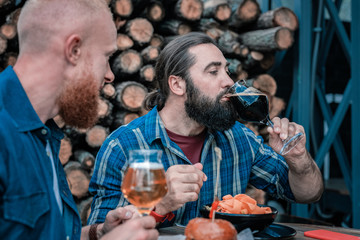 New taste. Mature dark-haired bearded man trying new taste of craft dark beer while interacting with his friend
