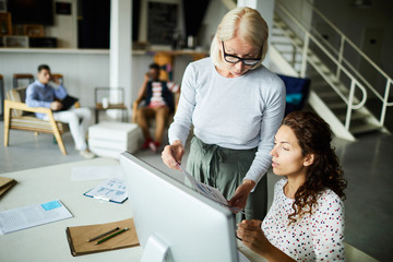 Obraz premium Mature female leader wearing eyeglasses discussing documents with businesswoman while she is sitting at the desk
