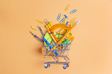 Shopping trolley with school supplies on yellow background. Flat lay, top view.