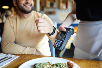 Smiling young man with smartwatch keeping his wrist close to payment machine while using transfer system to pay for meal