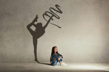 Baby girl dreaming about gymnast profession. Childhood and dream concept. Conceptual image with shadow of female gymnast on the studio wall - obrazy, fototapety, plakaty