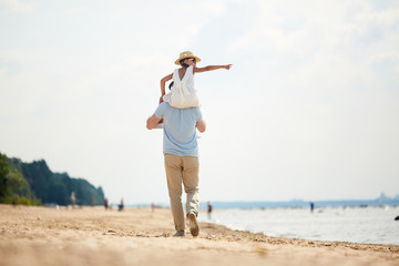 Back view of young man holding his little daughter on shoulders while walking down sandy beach at...