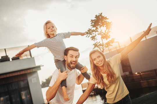 We can fly as planes. Portrait of optimistic bearded man keeping smiling girl on shoulders. She gesticulating hands with glad female. Happy parents and kid entertaining together outdoor concept