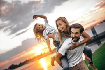 Portrait of beaming bearded dad keeping happy wife and outgoing daughter. Smiling family entertaining in evening outside concept