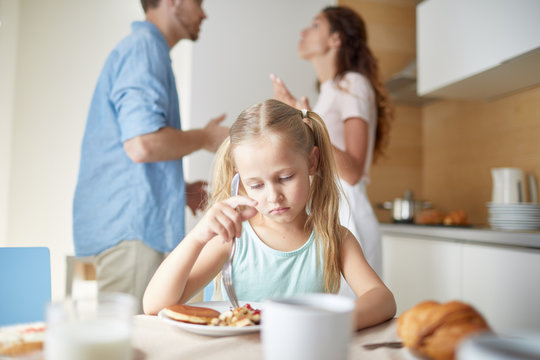 Upset and stressed little girl trying to eat while sitting by breakfast and her parents having argument on background