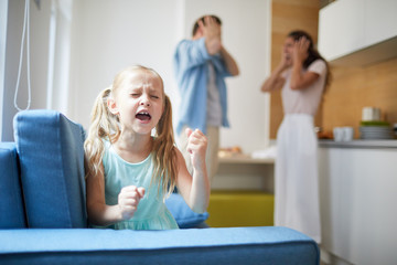 Little capricious girl expressing her displeasure by loud voice and hitting armchair with shocked parents on background - obrazy, fototapety, plakaty