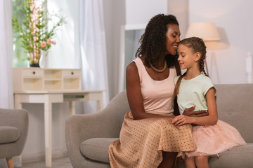 At home. Delighted positive woman sitting together with her daughter while hugging her