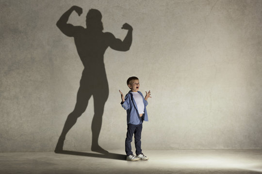 The little boy dreaming about athletic bodybuilder figure with muscles. Childhood and dream concept. Conceptual image with boy and shadow of fit athlete on the studio wall