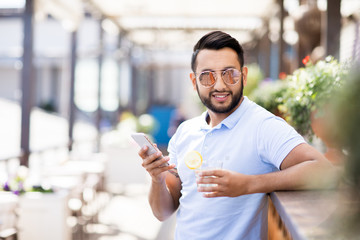 Young man in sunglasses and casualwear texting in smartphone and having drink in outdoor cafe