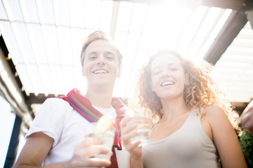 Young laughing couple with drinks looking at you on summer day while spending time in cafe
